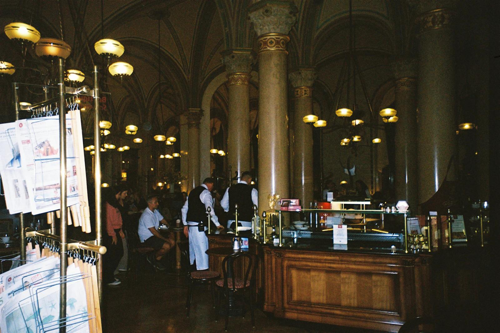 Café ornamentado e pouco iluminado, com colunas e pessoas às mesas, fotografado em Viena