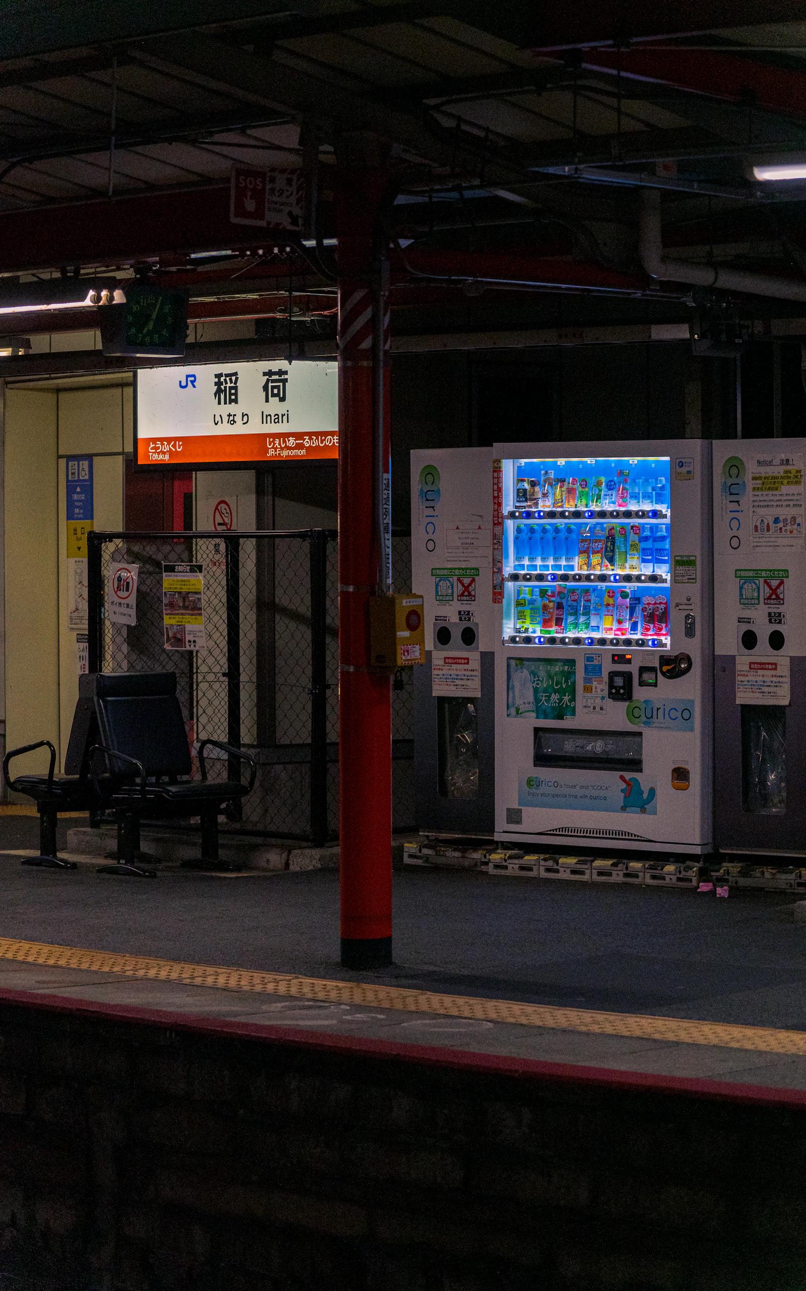 Plataforma de estação de trem à noite, com máquinas de venda acesas ao lado de um banco